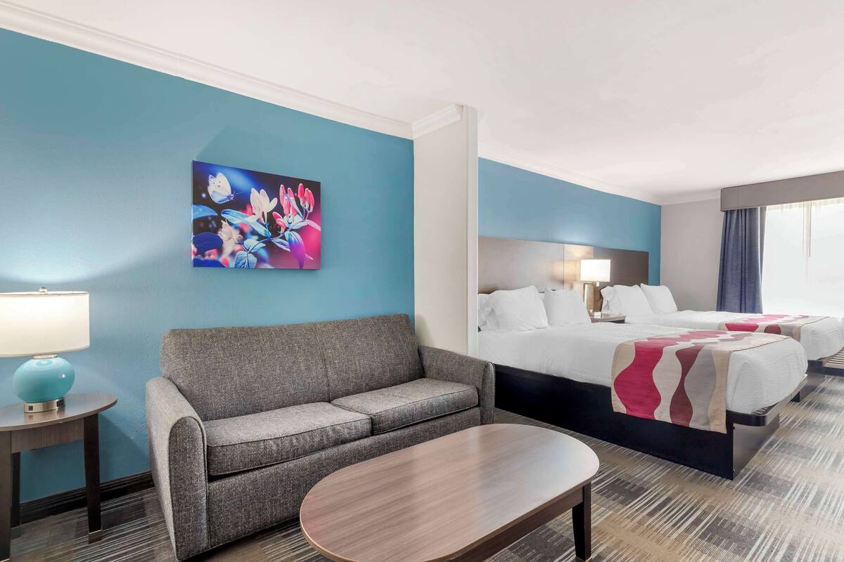Una camera d'albergo con un divano grigio, un tavolino in legno, un letto con biancheria bianca e un quadro appeso al muro.