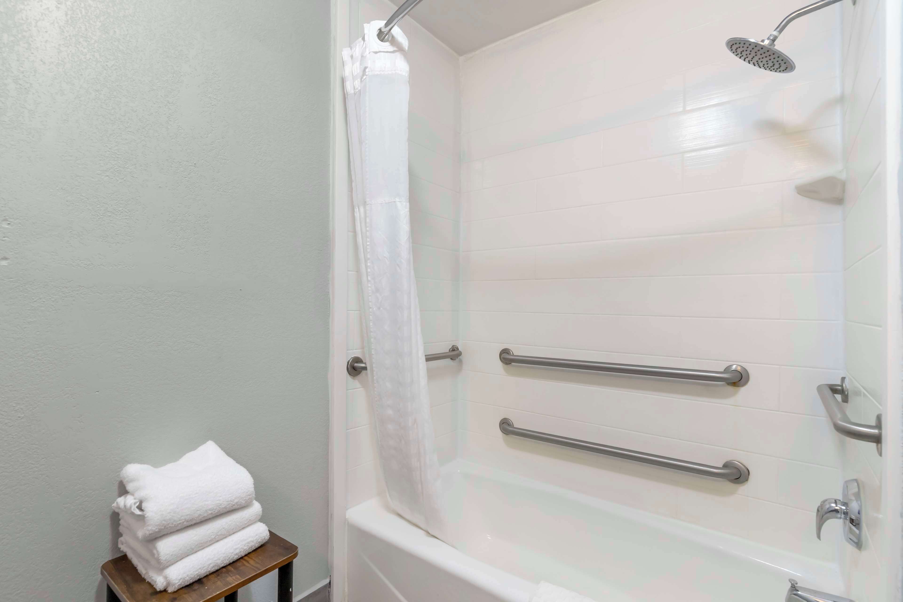 A bathroom with a white bathtub, a shower with a handheld showerhead, and a stack of white towels on a small stool.