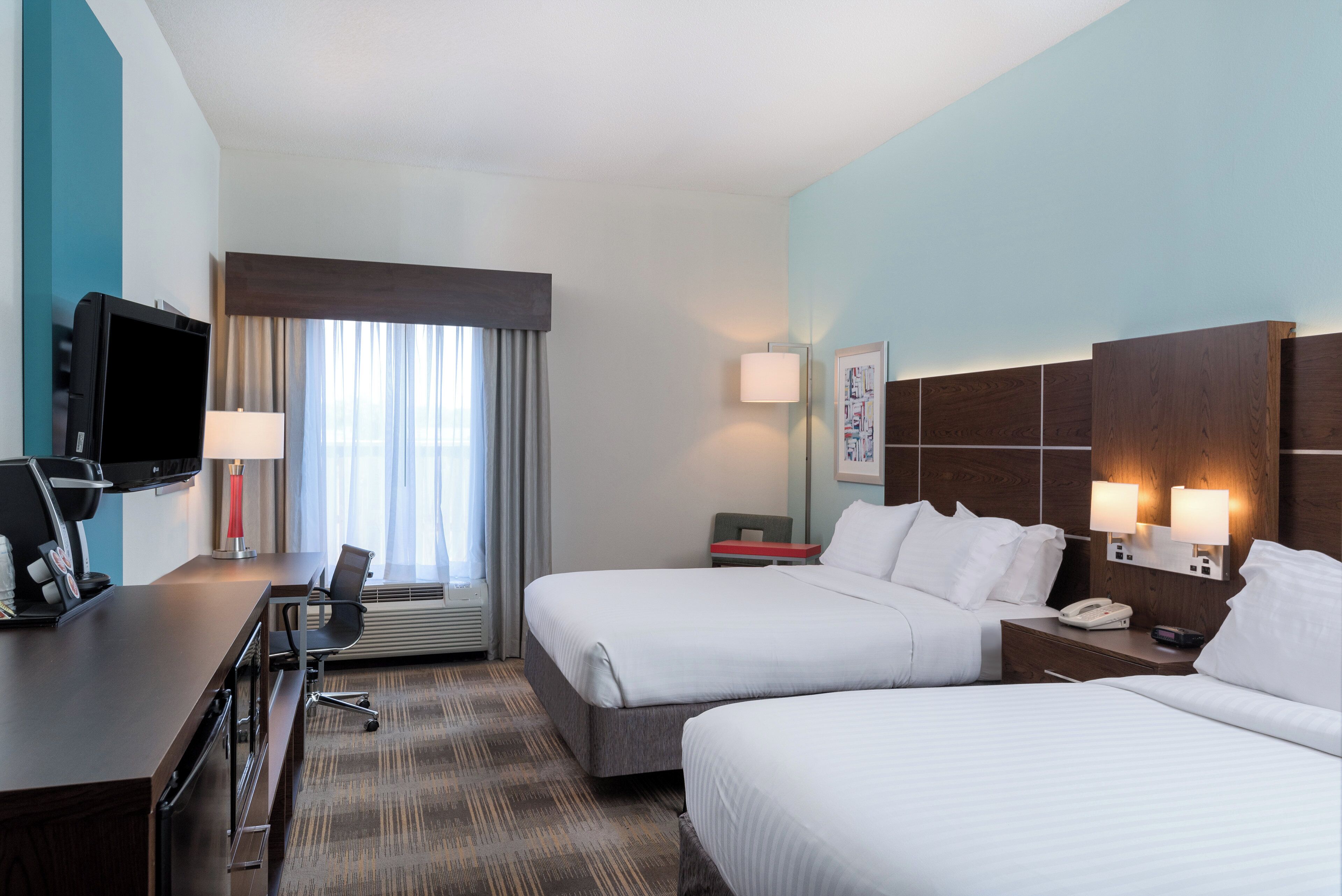 A hotel room with a bed, bedside table, lamp, TV, and a desk with a coffee machine.