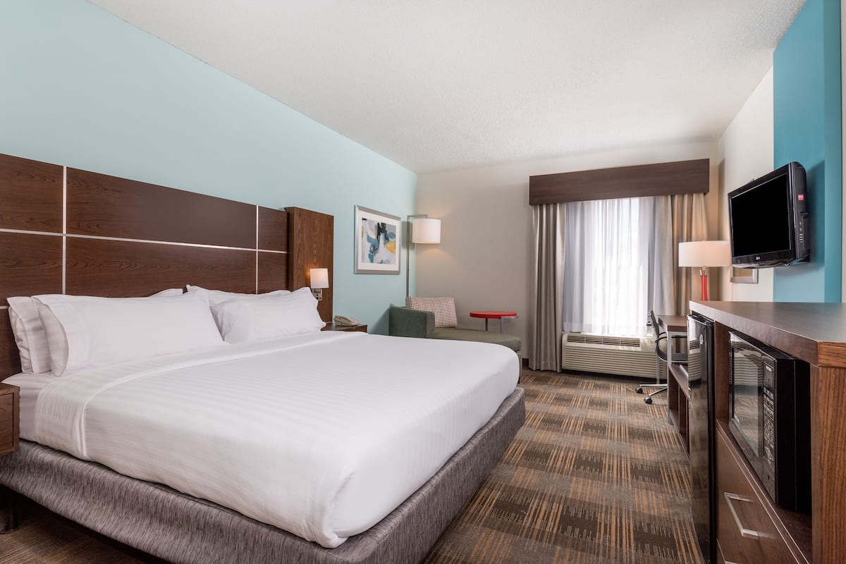 A hotel room with a large bed, a desk with a flat-screen TV, a chair, a small table, and a window with curtains.