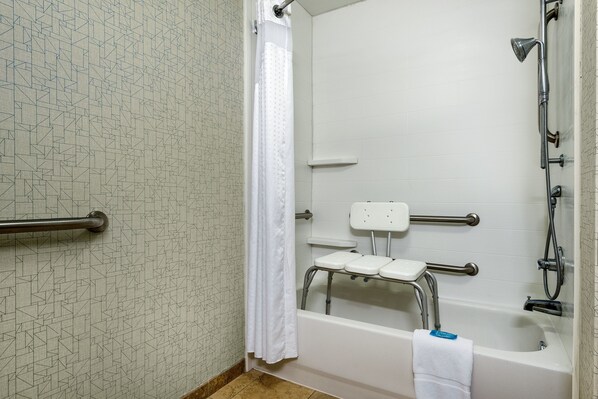 A bathroom with a shower, grab bars, a wheelchair-accessible bench, and a white tiled wall.