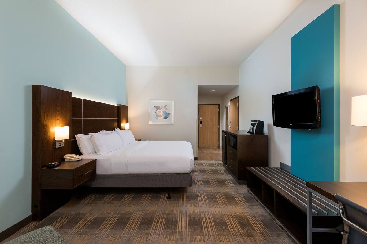 A hotel room with a bed, bedside table with a lamp, a TV mounted on the wall, and a bench.