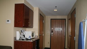 A hotel room with a kitchenette, a microwave, a coffee maker, and a small refrigerator. There is a door with a sign on it, and a coat rack with a blue item hanging on it. - Holiday Inn Express & Suites Grenada by IHG (Grenada)