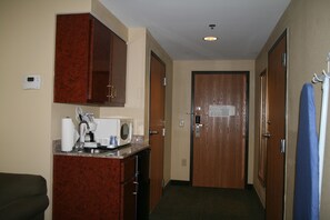 A hotel room with a kitchenette, a microwave, a coffee maker, and a small refrigerator. There is a door with a sign on it, and a coat rack with a blue item hanging on it. - Holiday Inn Express & Suites Grenada by IHG (Grenada)
