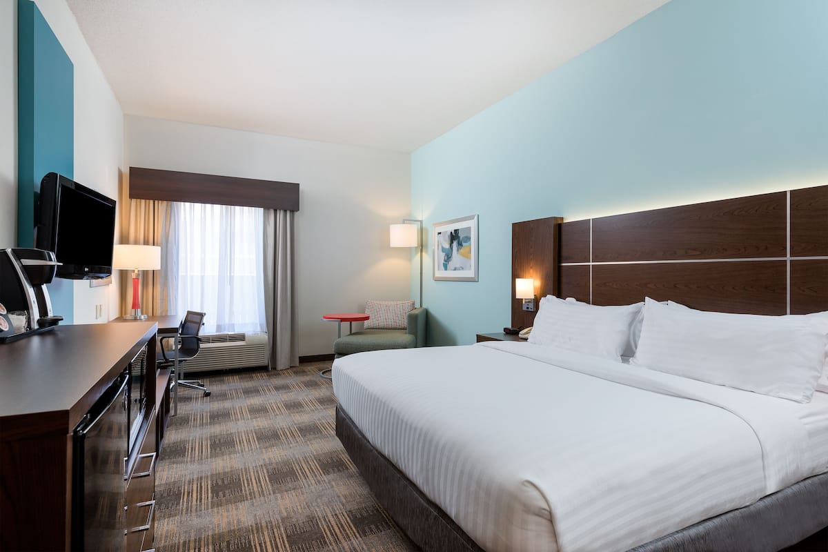 A hotel room with a large bed, a desk with a TV, a chair, a small table, and a lamp.