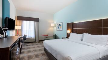 Quarto de hotel com uma cama grande, uma escrivaninha com televisão, uma cadeira, uma mesa pequena e um abajur.