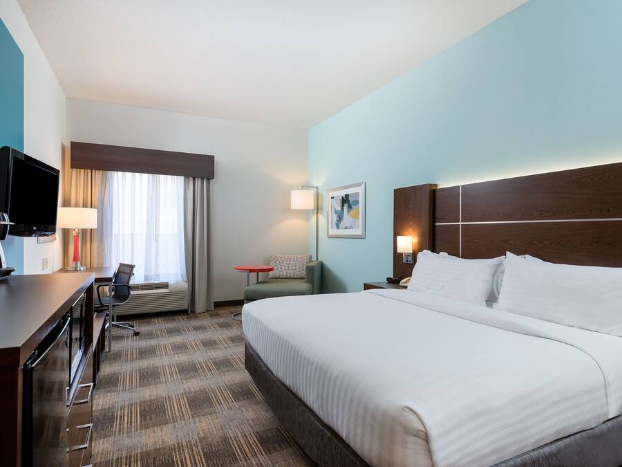 Quarto de hotel com uma cama grande, uma escrivaninha com televisão, uma cadeira, uma mesa pequena e um abajur.