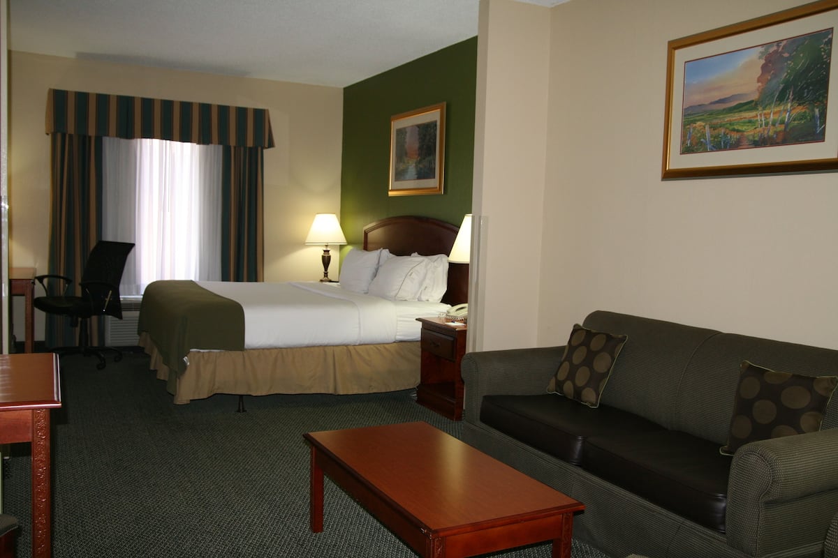 A hotel room with a bed, a wooden nightstand, a desk, a chair, a sofa, and a painting on the wall.