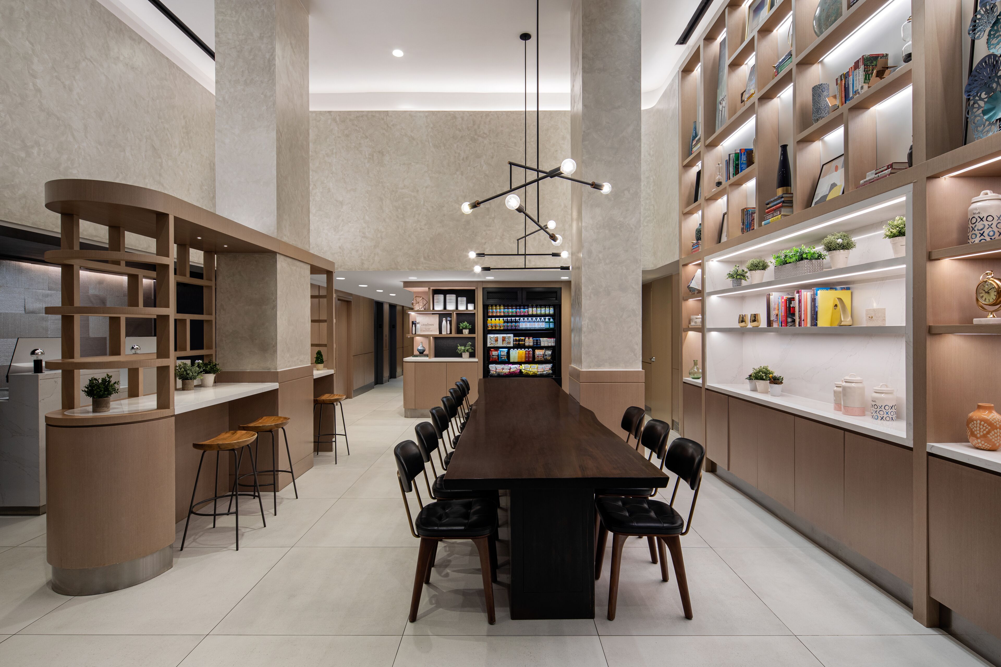 Un espace de restauration moderne et bien éclairé, avec une longue table, des tabourets de bar et des étagères remplies de livres et d’objets décoratifs.