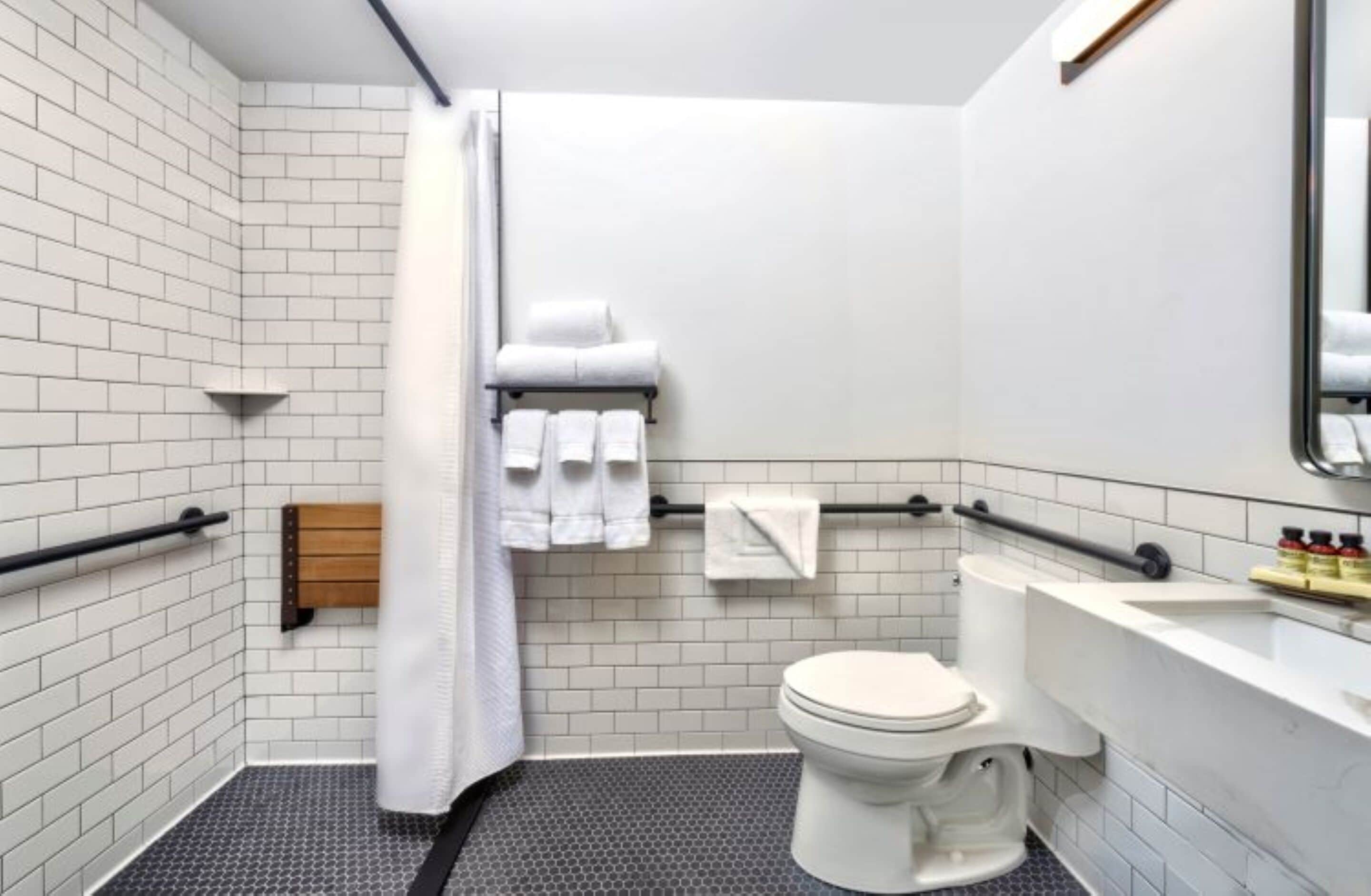 Une salle de bain moderne avec des carreaux de céramique blancs rectangulaires, des toilettes blanches, une douche avec une porte en verre et un porte-serviettes avec plusieurs serviettes.