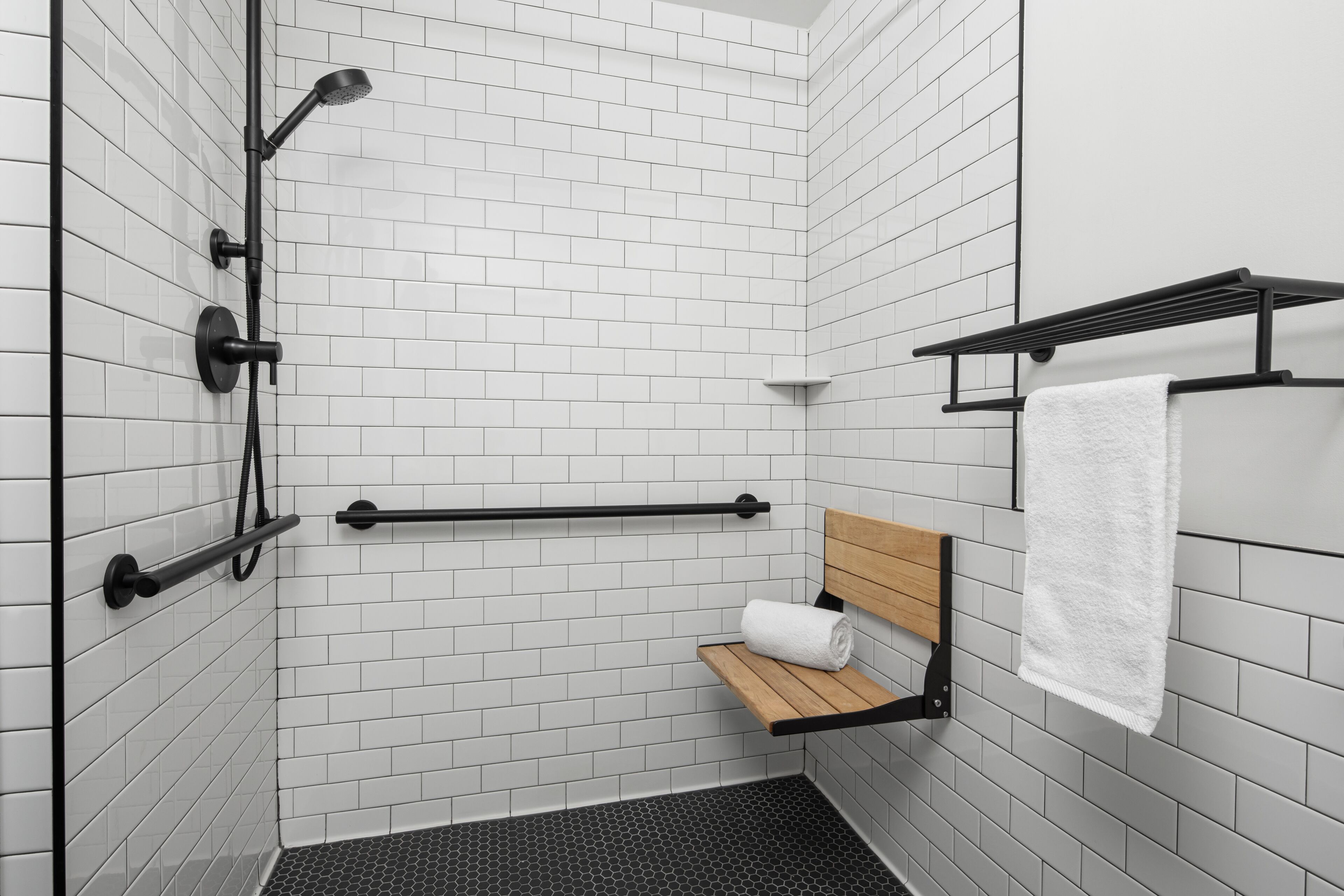 A modern bathroom with white tiled walls, a shower with black fixtures, a wooden bench, and a towel rack.