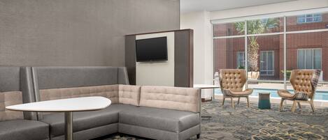 A modern hotel room with a gray sectional sofa, a small white table, and two leather armchairs. A flat-screen TV is mounted on the wall, and a large window offers a view of a brick building and a pool.