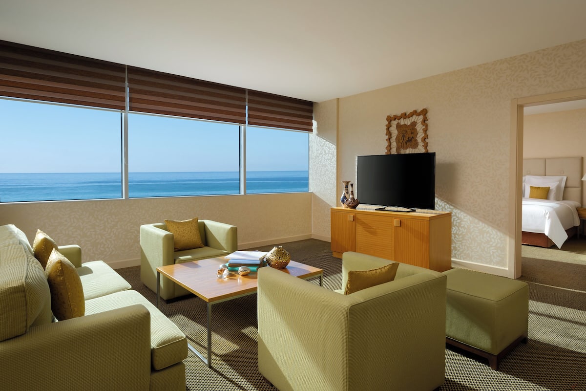 Ein Hotelzimmer mit großem Fenster und Meerblick, einem Fernseher auf einer Holzkabine, einem Sofa, einem kleinen Tisch und einem Bett mit Nachtschränkchen.