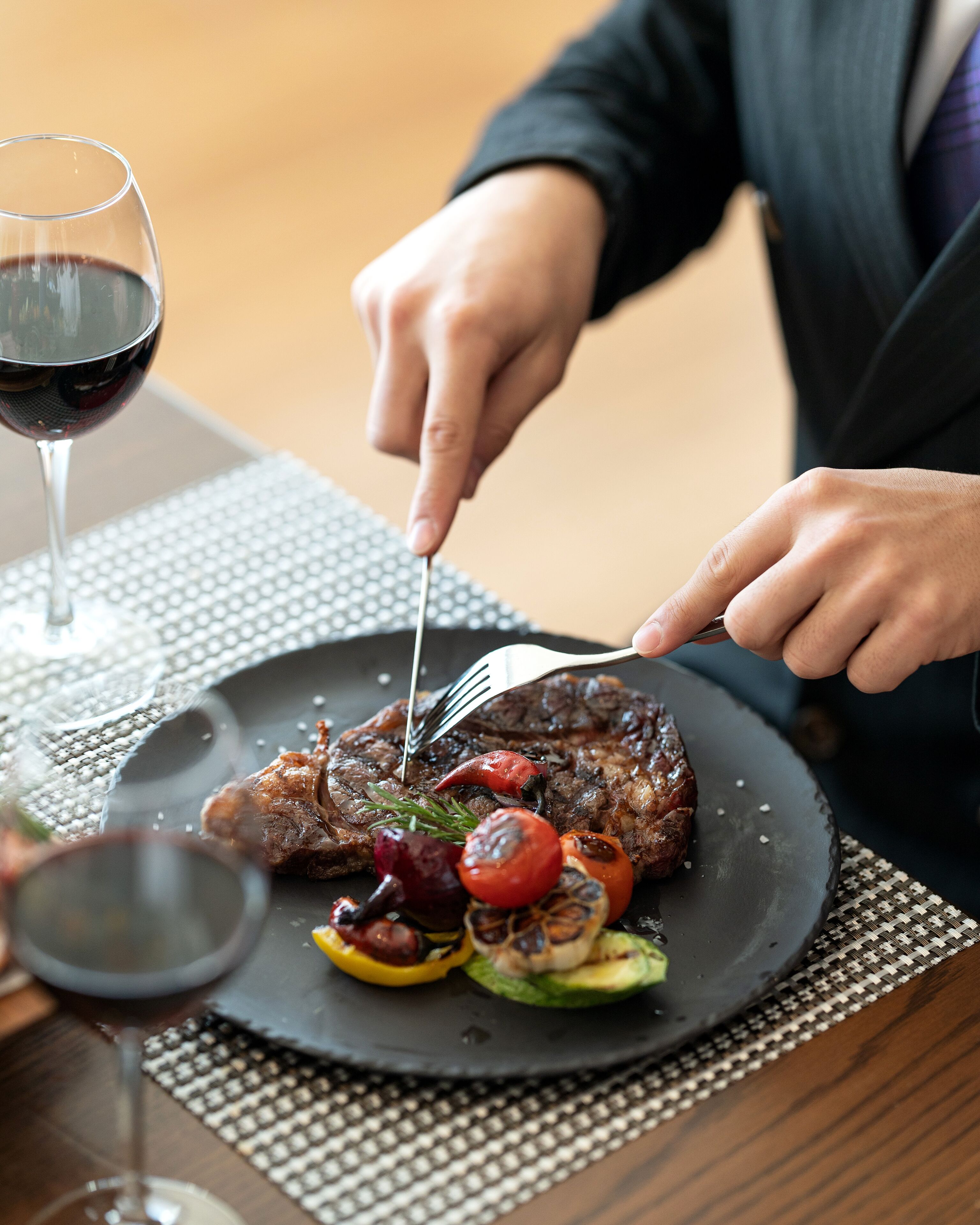 A steak with vegetables on a plate, a glass of red wine, and a person cutting the steak.