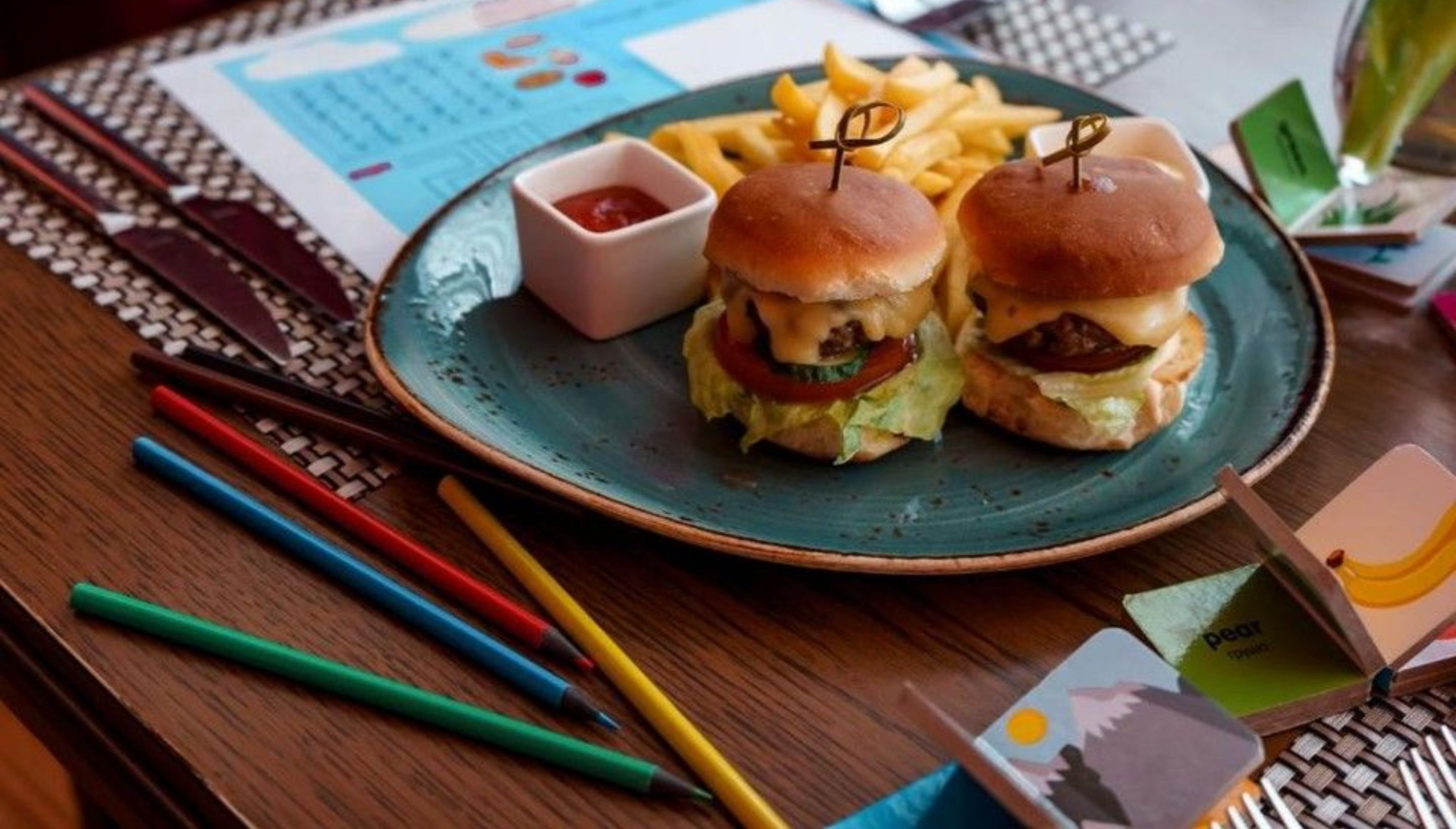 A plate with two burgers and fries, a small bowl of ketchup, and colored pencils on a wooden table.