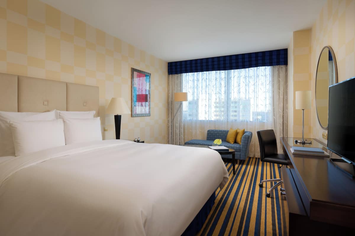 Ein Hotelzimmer mit einem großen Bett, einem Schreibtisch mit Stuhl, einem Fernseher und einem Fenster mit Vorhängen.