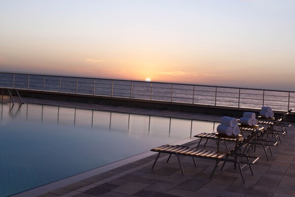 A poolside area with lounge chairs and sun loungers, a railing, and a sunset view.
