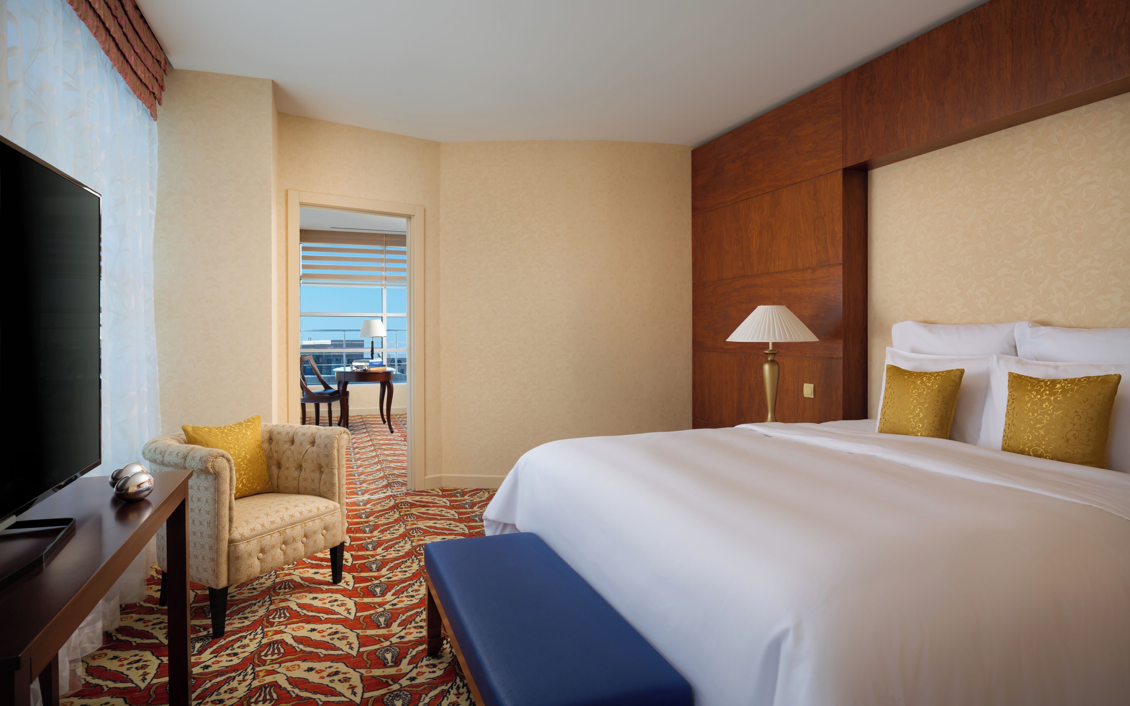 A hotel room with a large bed, a desk with a TV, a chair, a lamp, and a view of the city through the window.