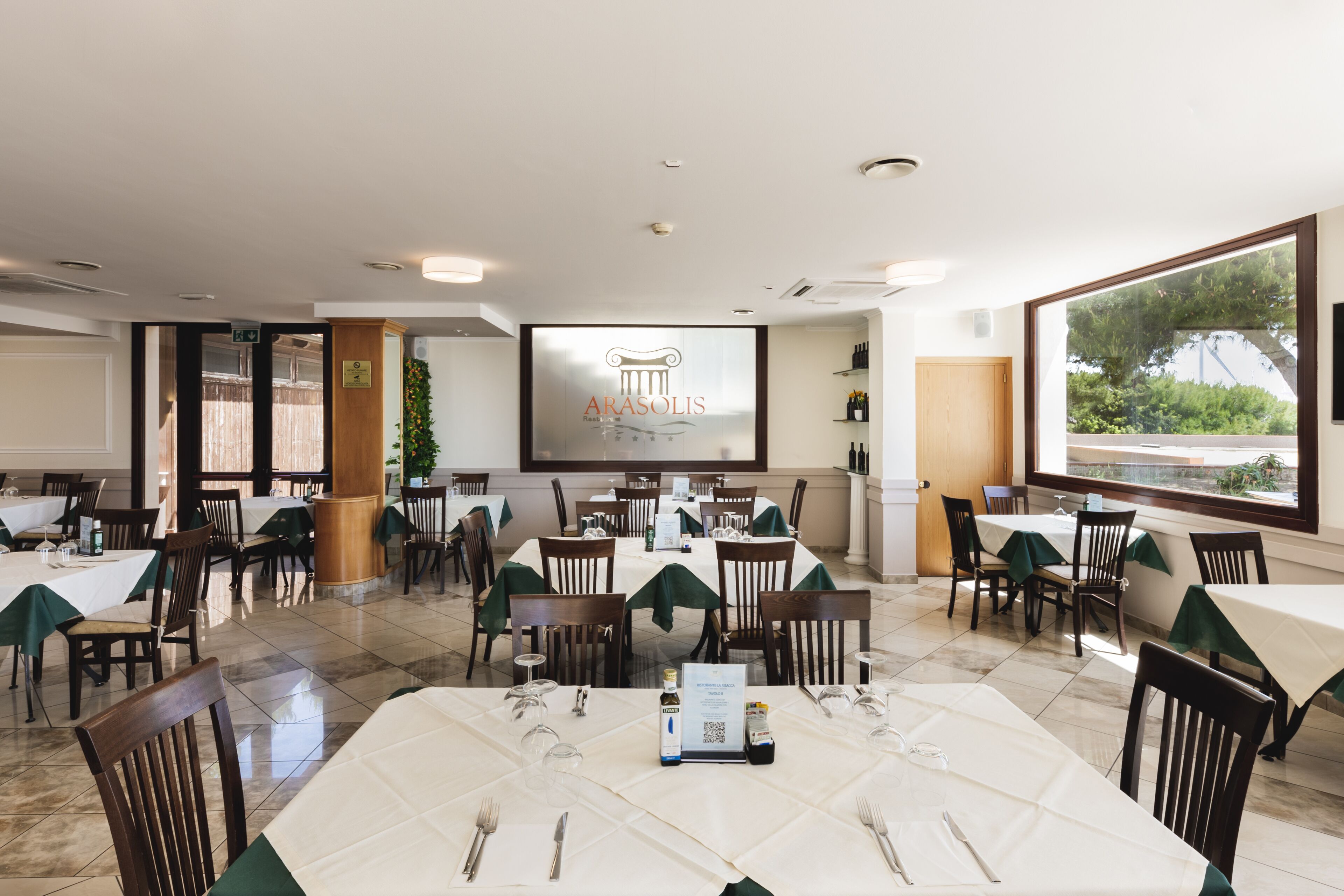 A restaurant with multiple tables and chairs, a large screen displaying the restaurant's name, and a view of the outdoors through large windows.
