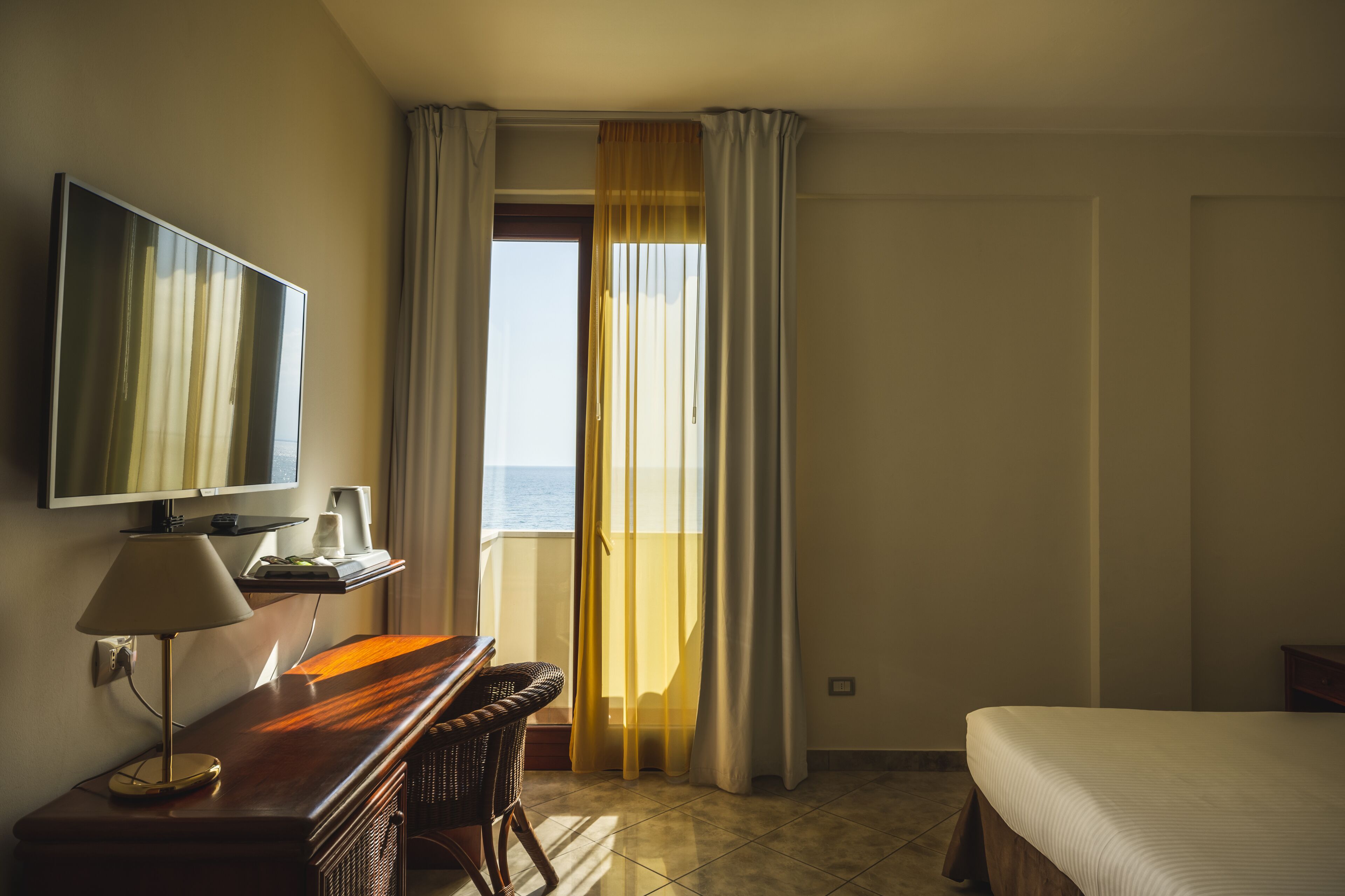 A hotel room with a bed, a desk with a lamp, a television, and a view of the sea through a window with curtains.