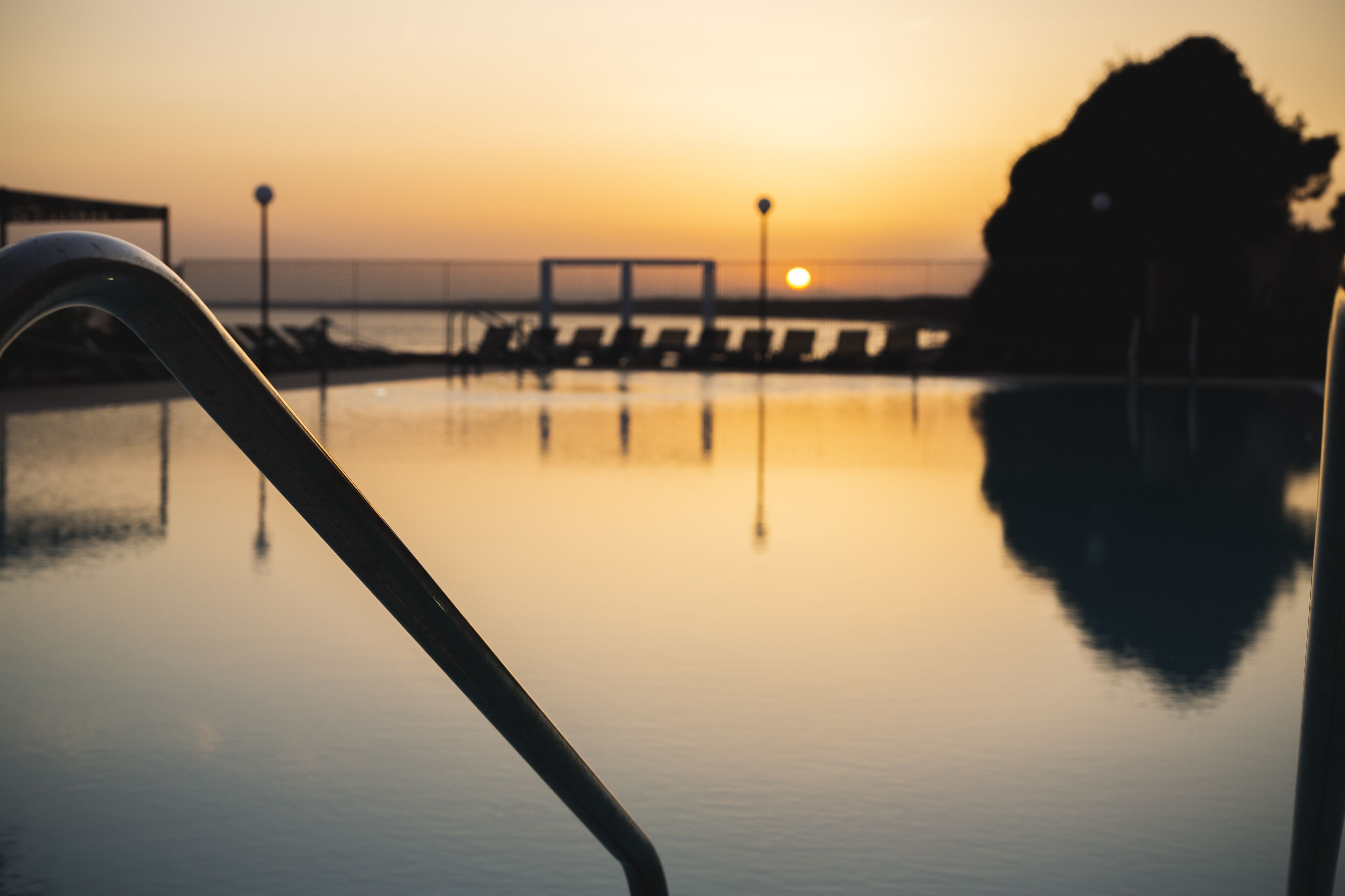 Un'area piscina tranquilla con vista al tramonto, una ringhiera in metallo e un molo in lontananza.