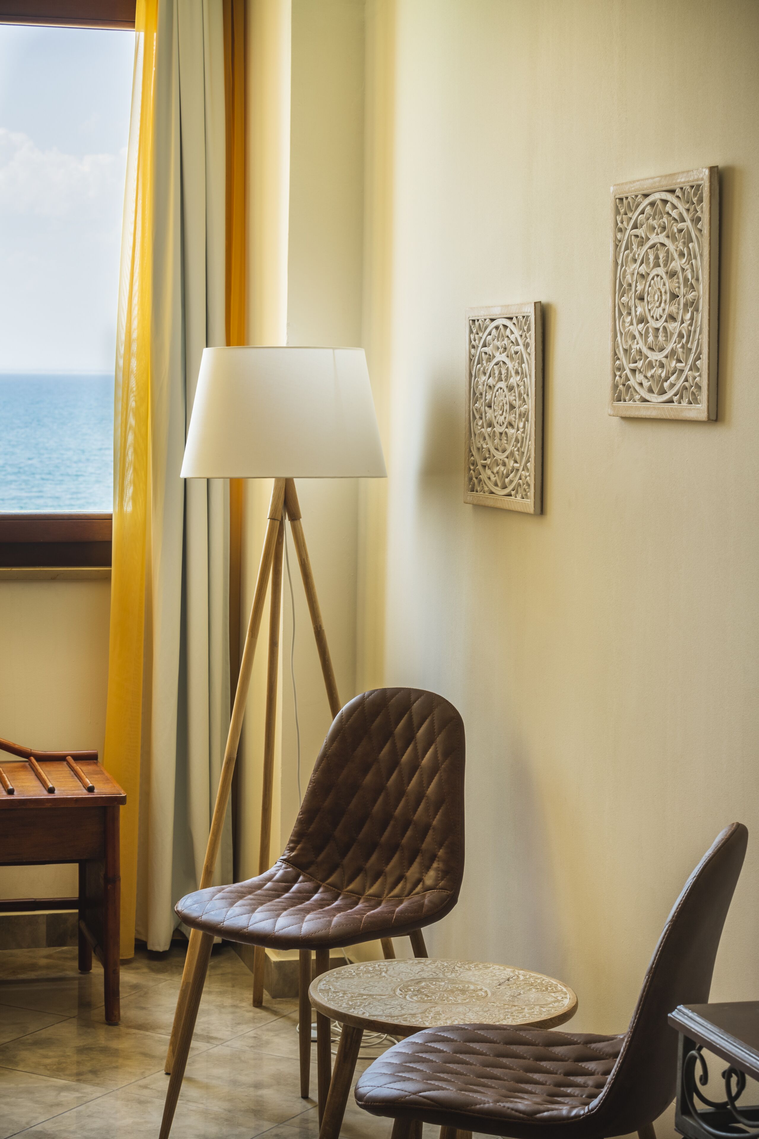 A room with a view of the sea, a lamp, two chairs, a small table, and decorative wall art.