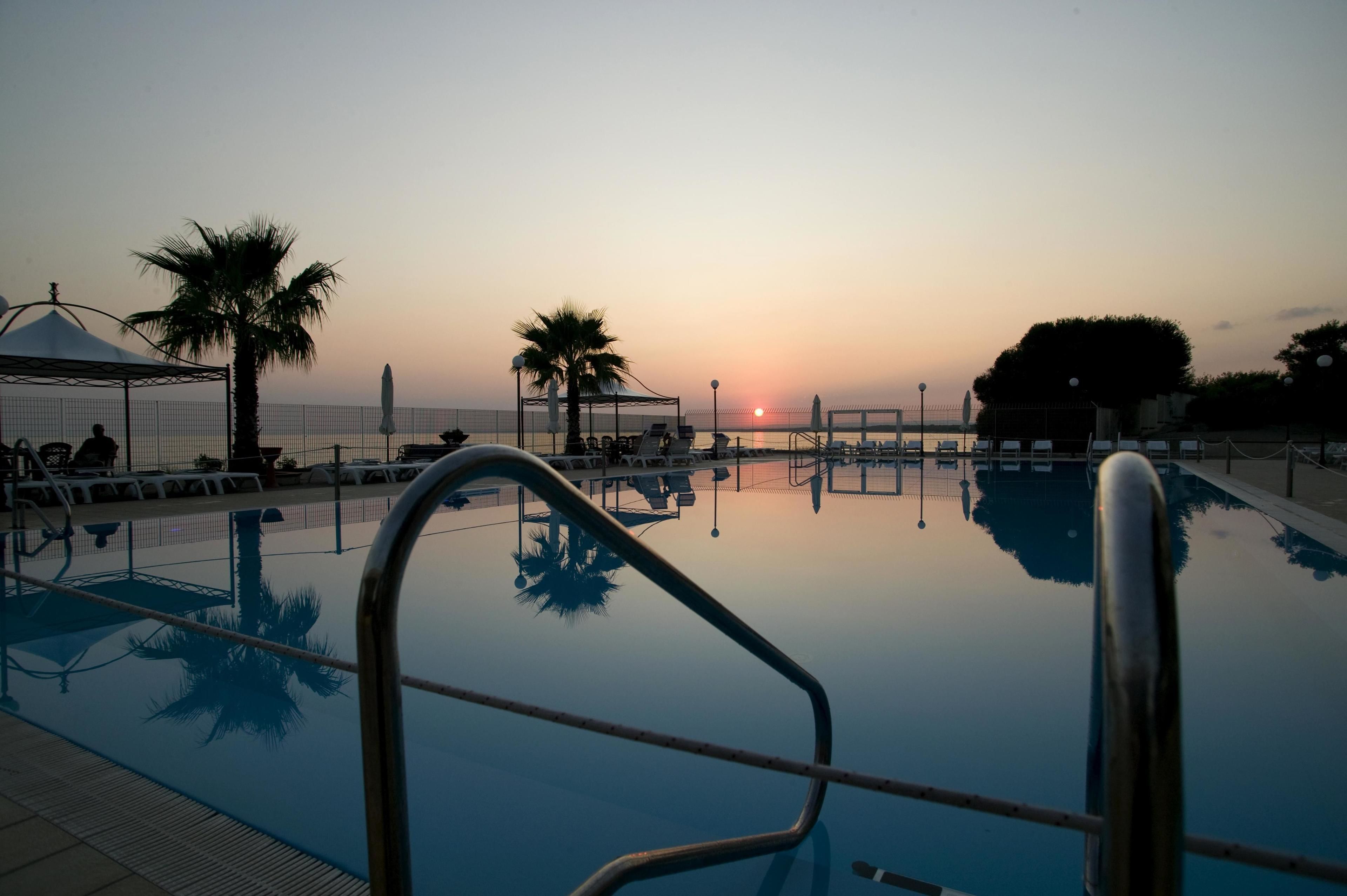 A swimming pool with a clear view of the sunset, palm trees, and a covered seating area.