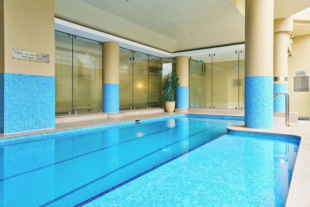 An indoor swimming pool with clear glass walls, blue-tiled steps, and a sign indicating the pool is 12 meters deep.