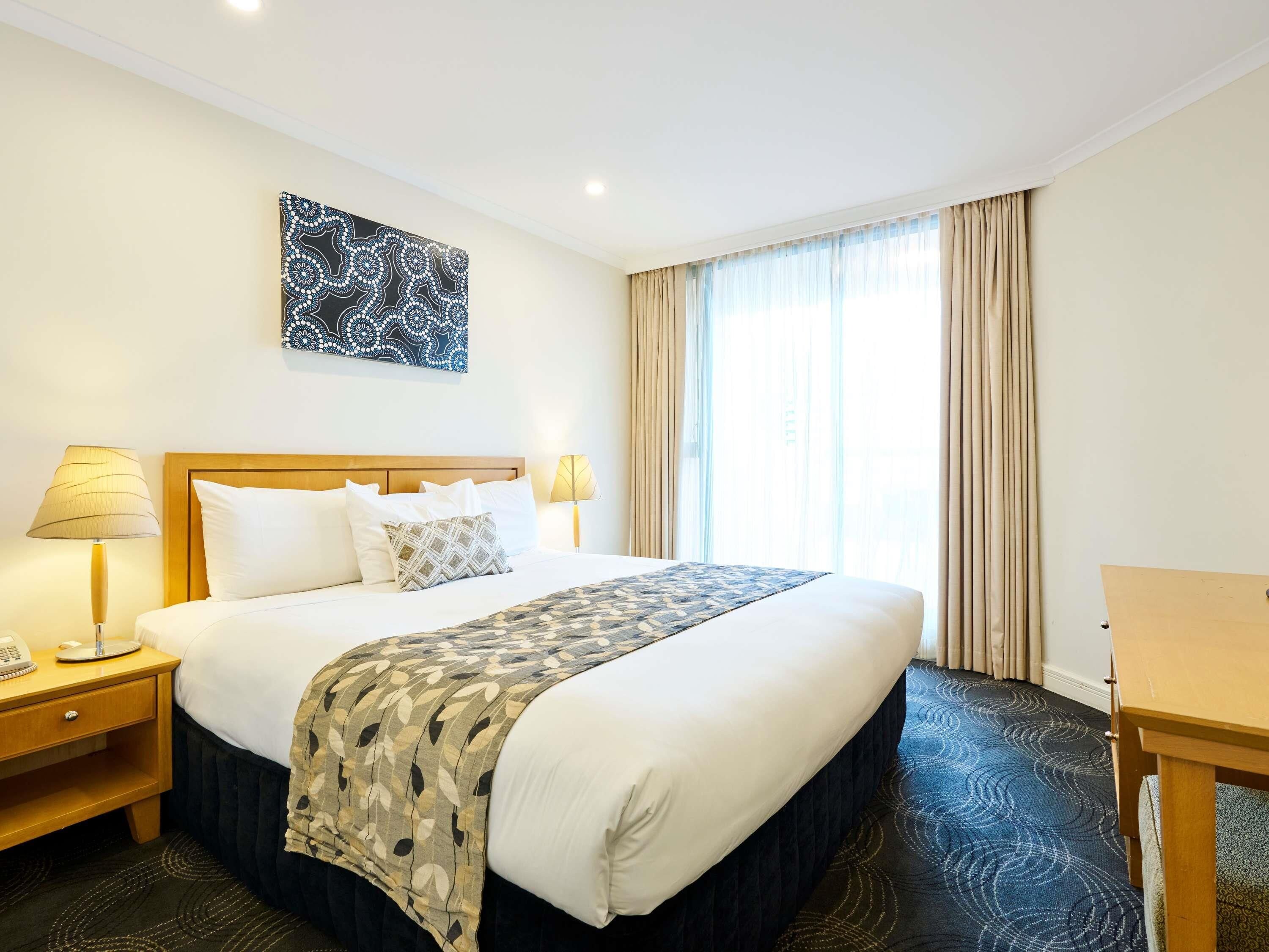 A hotel room with a large bed, two bedside lamps, a telephone, a desk, and a window with curtains.