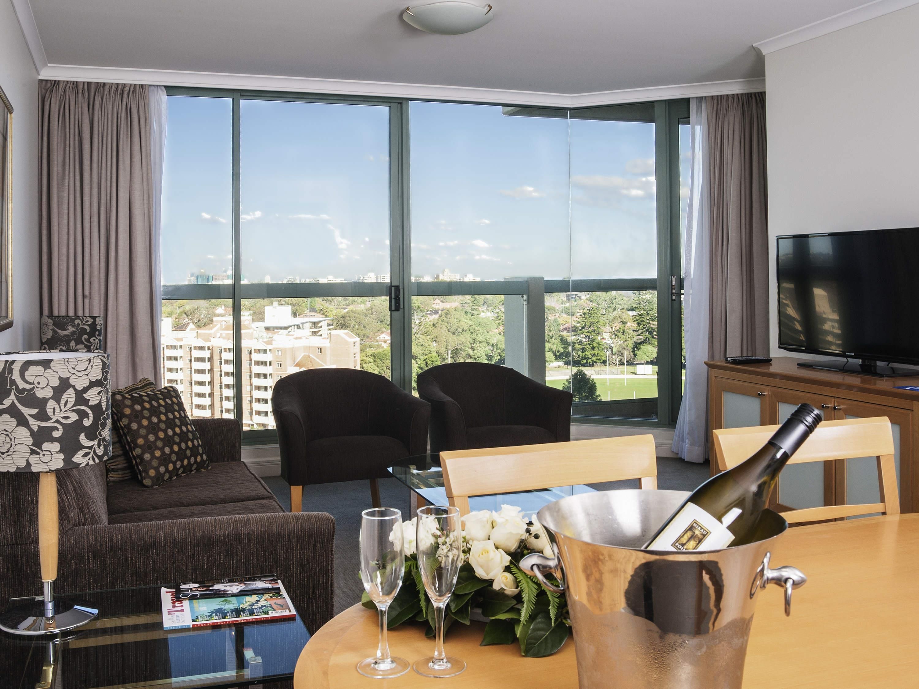 A hotel room with a large window, a television, a dining table with a bottle of champagne, two chairs, a sofa, and a magazine on the table.