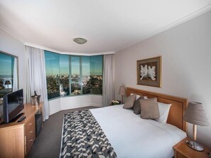 A hotel room with a large bed, a desk with a TV, a lamp, and a view of a cityscape through large windows.
