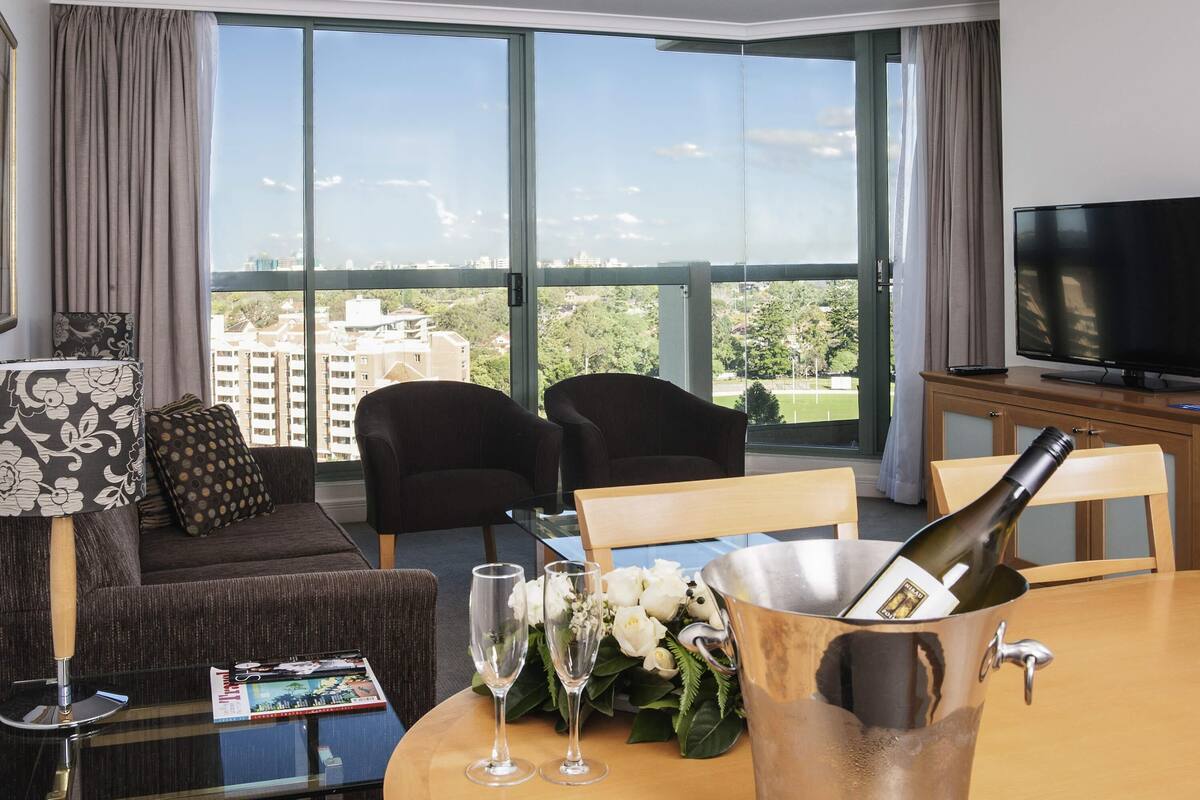 A hotel room with a large window, a television, a dining table with a bottle of champagne, two chairs, a sofa, and a magazine on the table.