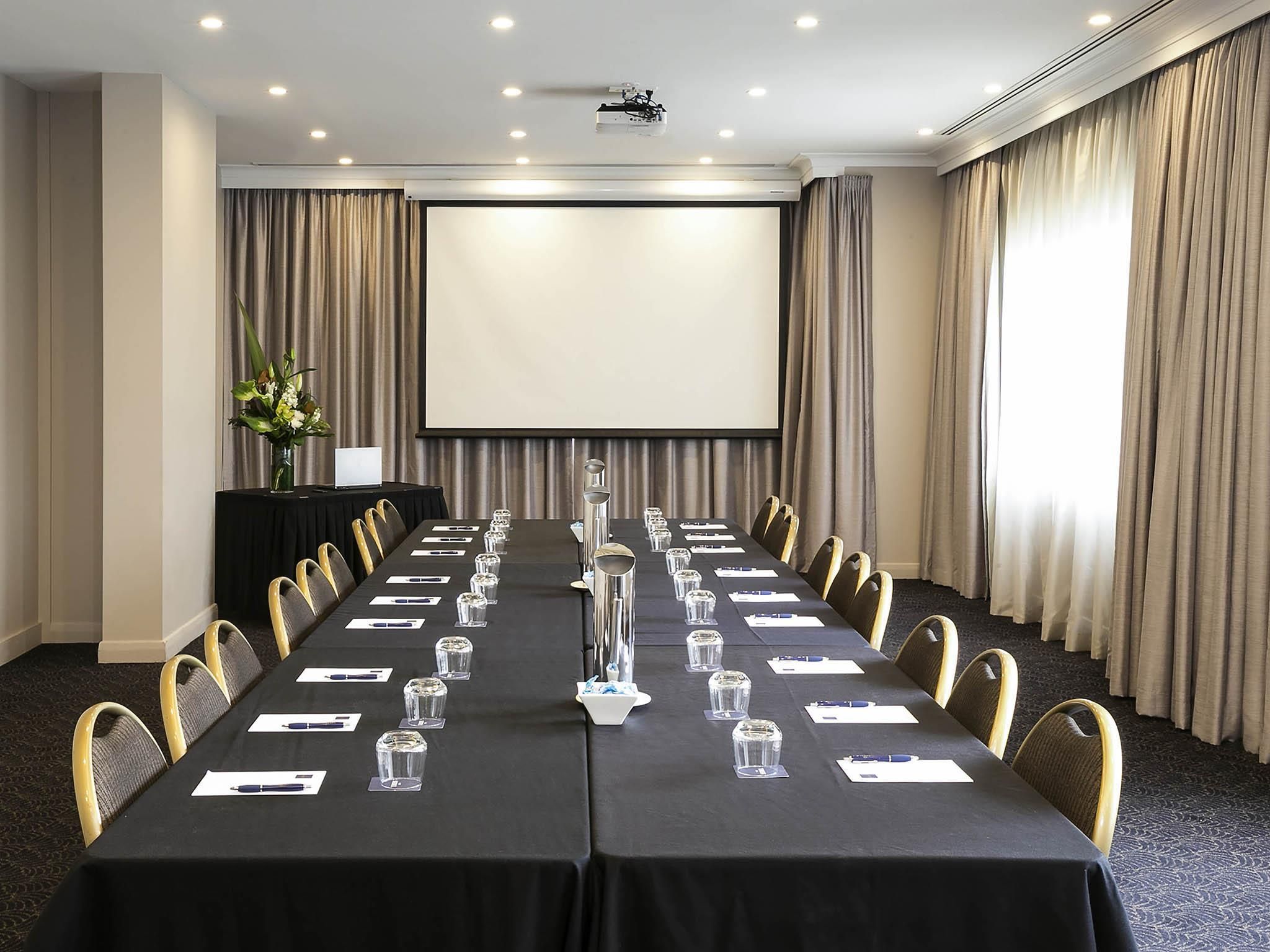 Un salón de conferencias con una larga mesa, sillas, una pantalla de proyector y cortinas.
