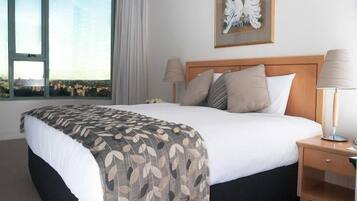 Quarto de hotel com uma cama grande, mesa de cabeceira com abajur, janela com vista e uma obra de arte emoldurada na parede.