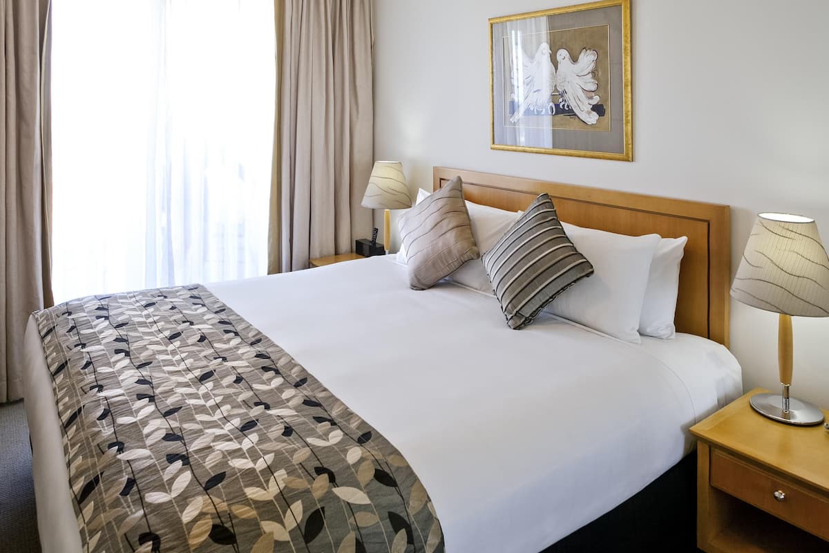 A hotel room with a neatly made bed, a wooden headboard, a bedside table with a lamp, and a framed artwork on the wall.