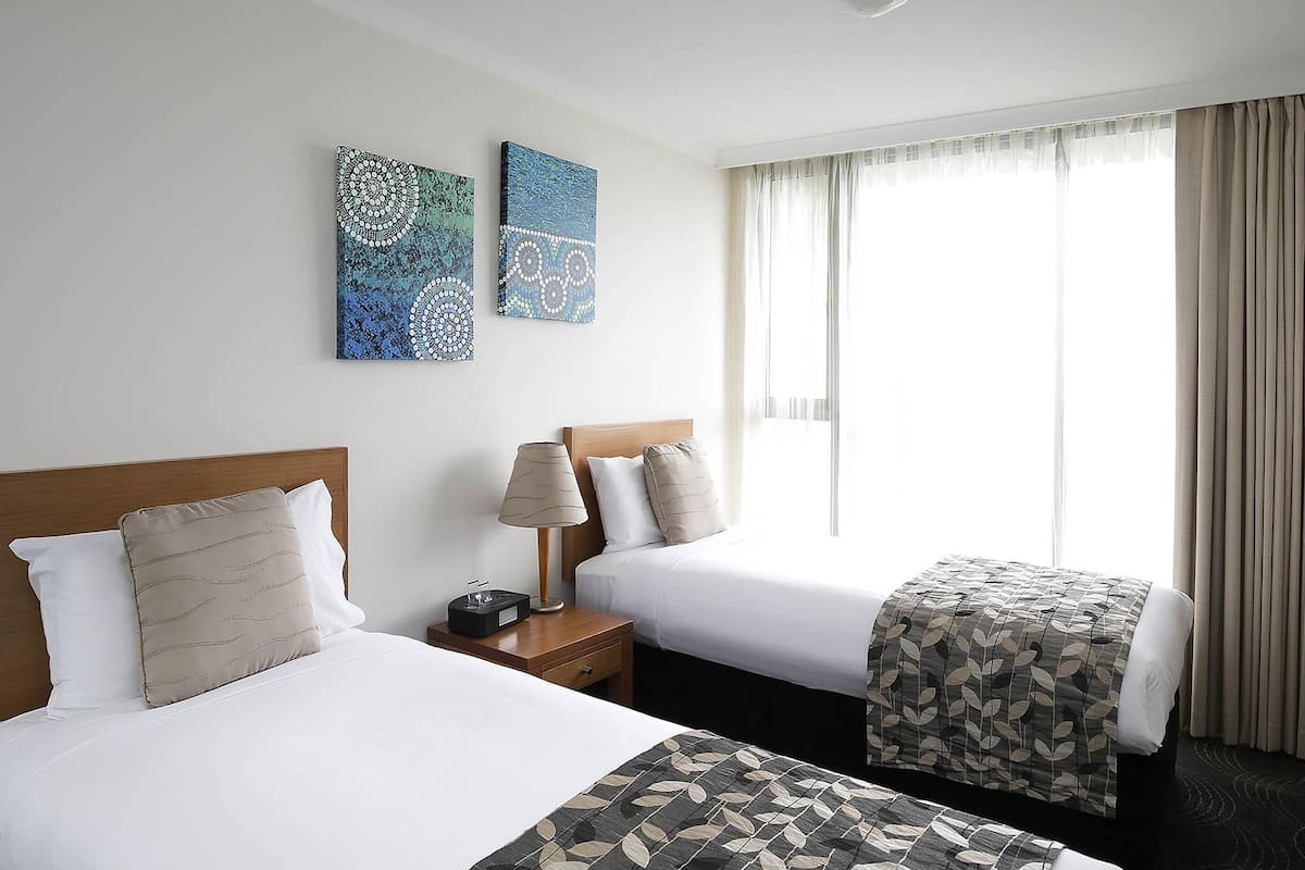A hotel room with two beds, a nightstand with a lamp, and artwork on the wall.