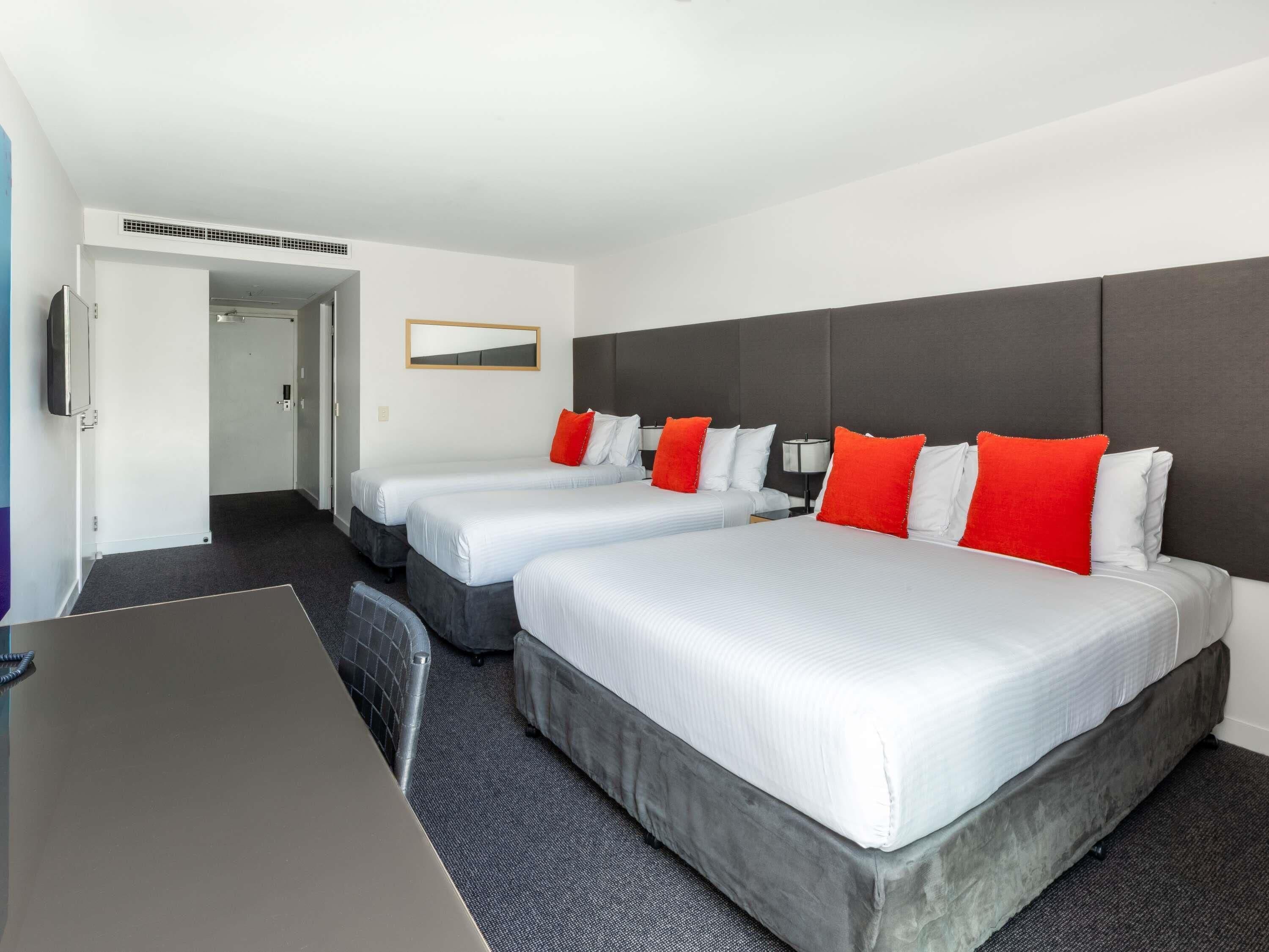 Ein Hotelzimmer mit drei Betten, einem Schreibtisch, einem Fernseher und einem Spiegel.