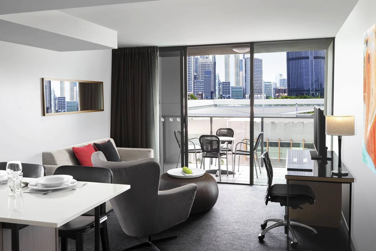 A modern hotel room with a sofa, dining table, chairs, a TV, and a balcony with a city view.