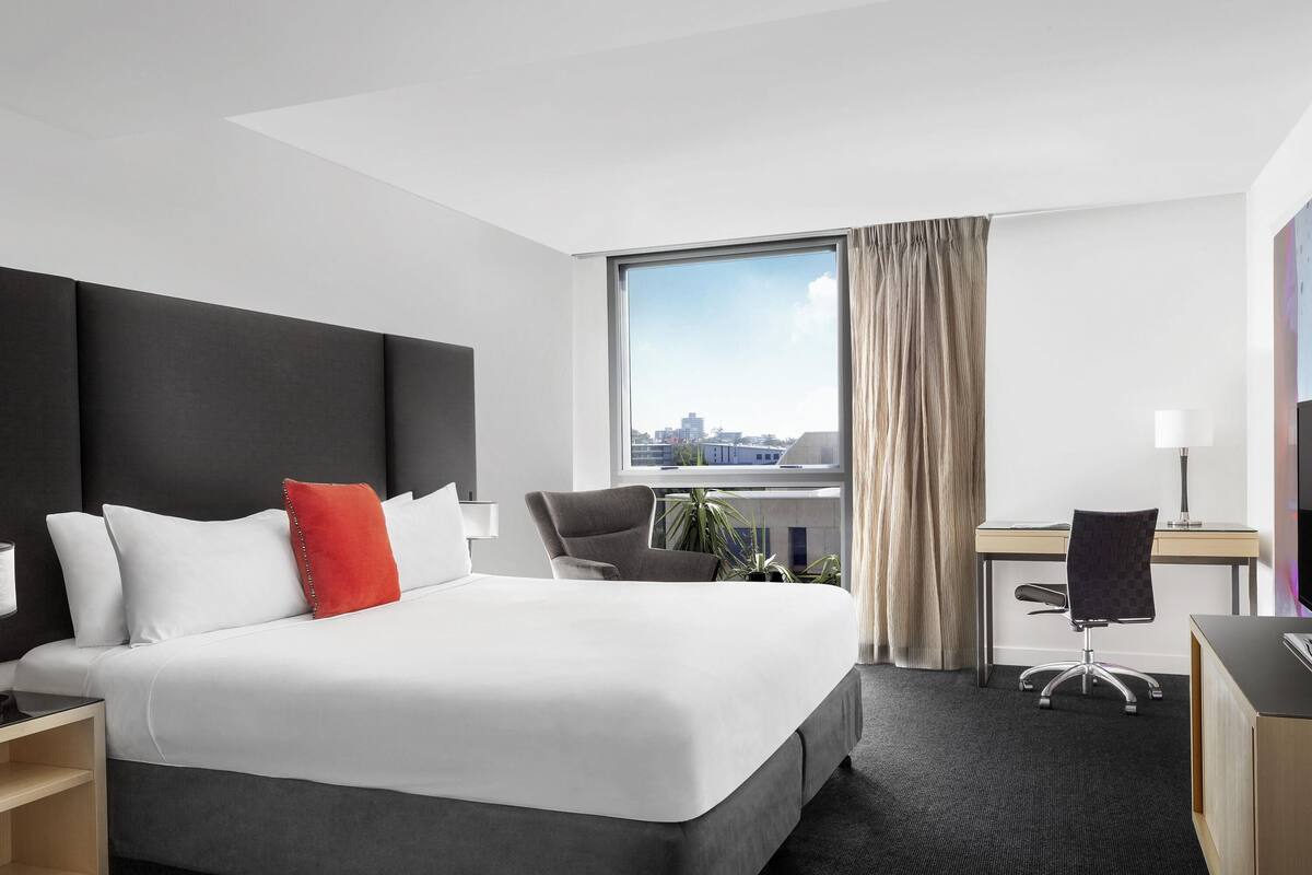 A hotel room with a large bed, a desk, a chair, a TV, and a window with a city view.