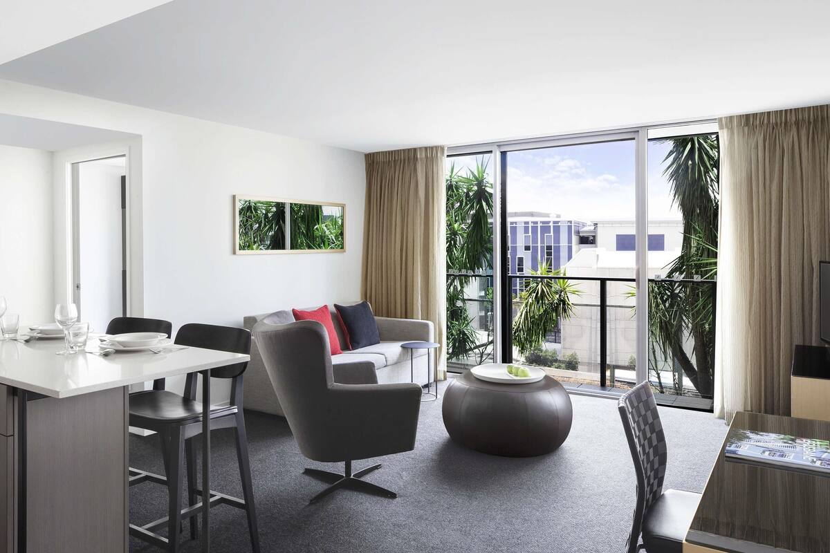 A modern hotel room with a dining area, a sofa, a coffee table, and large windows with a view of buildings and greenery.