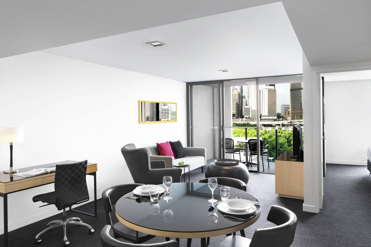 A modern hotel room with a dining area, a sofa, and a view of the cityscape.