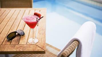 A wooden table with a cocktail, sunglasses, and a wicker chair by a pool.