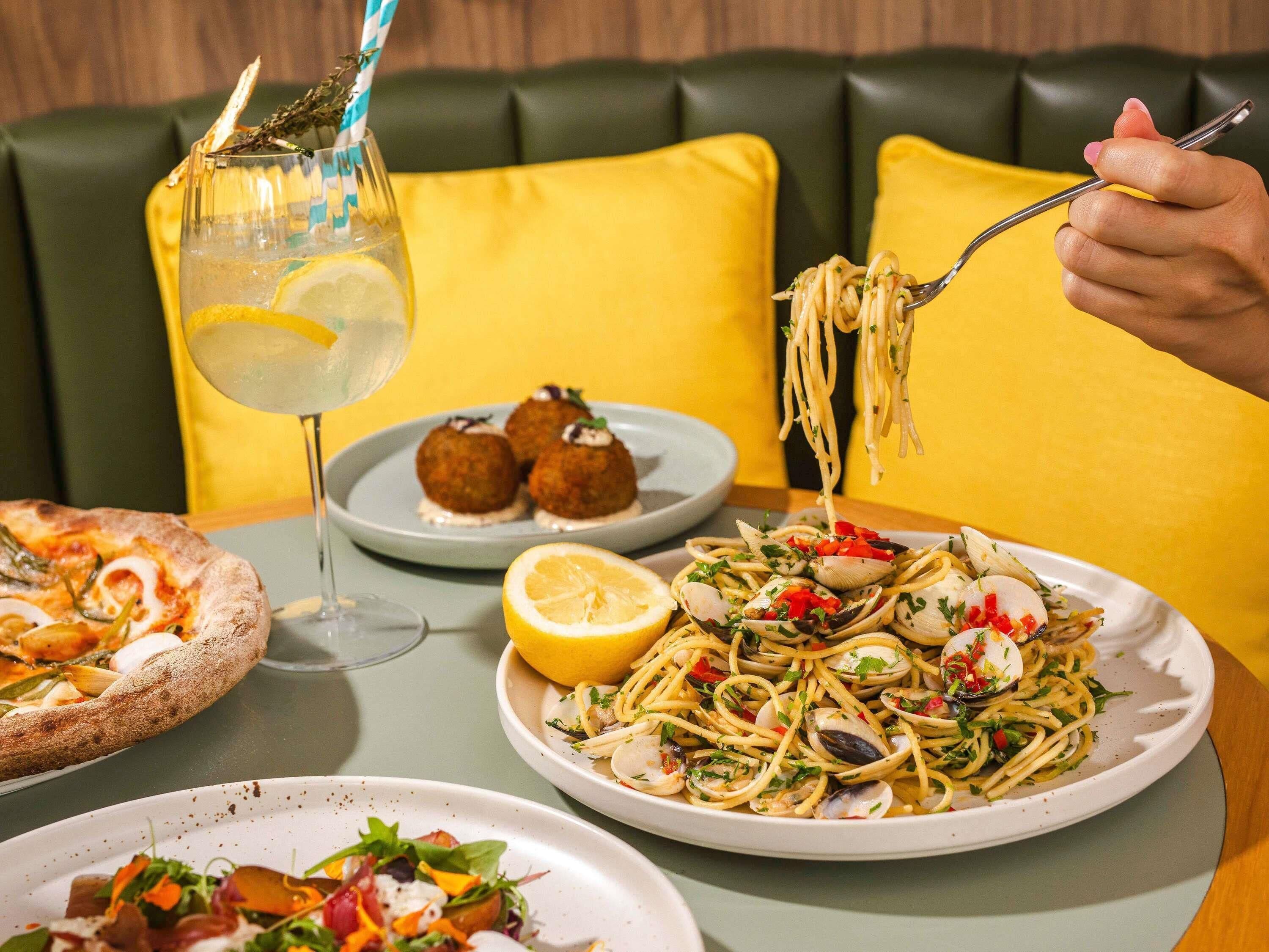 A dining table with plates of food, a glass of drink, and a person eating spaghetti.
