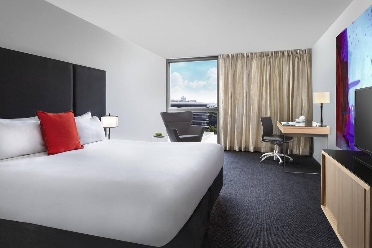 A hotel room with a large bed, a desk with a chair, a television, and a view of the city through the window.