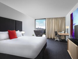 A hotel room with a large bed, a desk with a chair, a television, and a view of the city through the window.