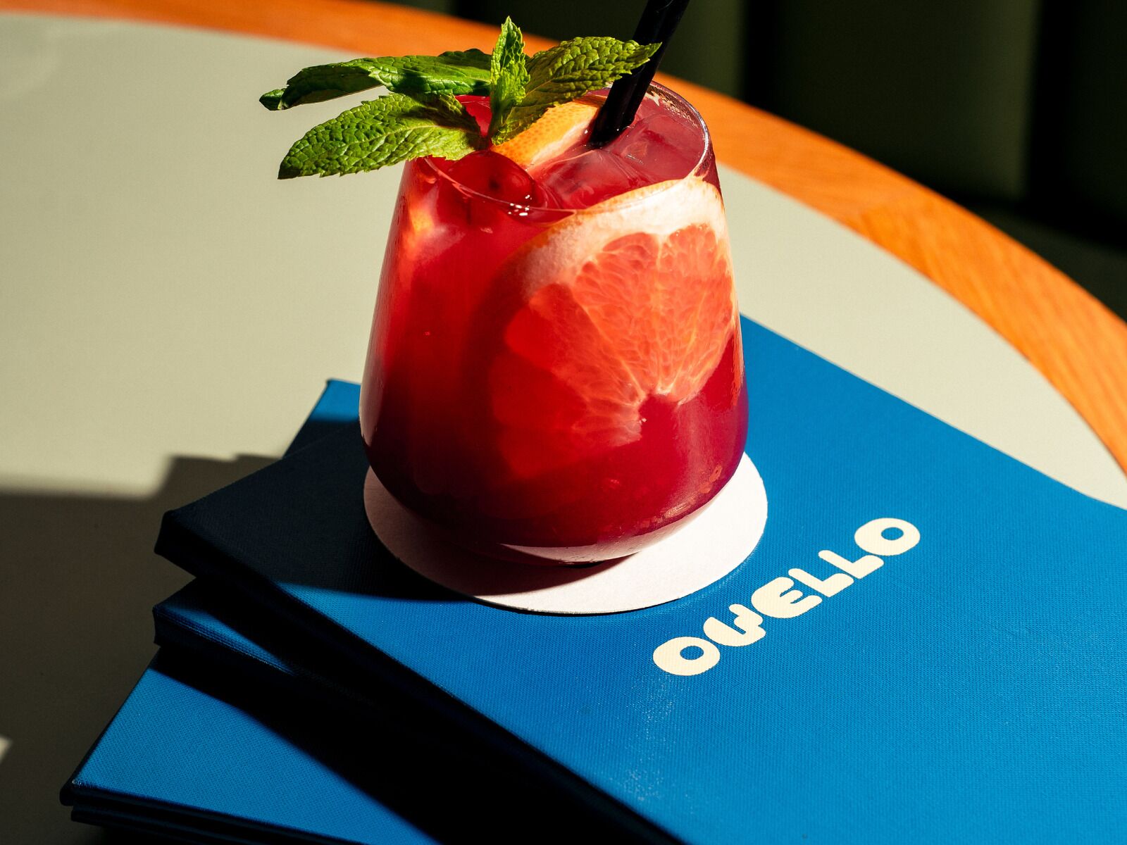 A glass of red beverage with a slice of fruit and mint garnish on a blue napkin with the word 'OVELLO' on it.