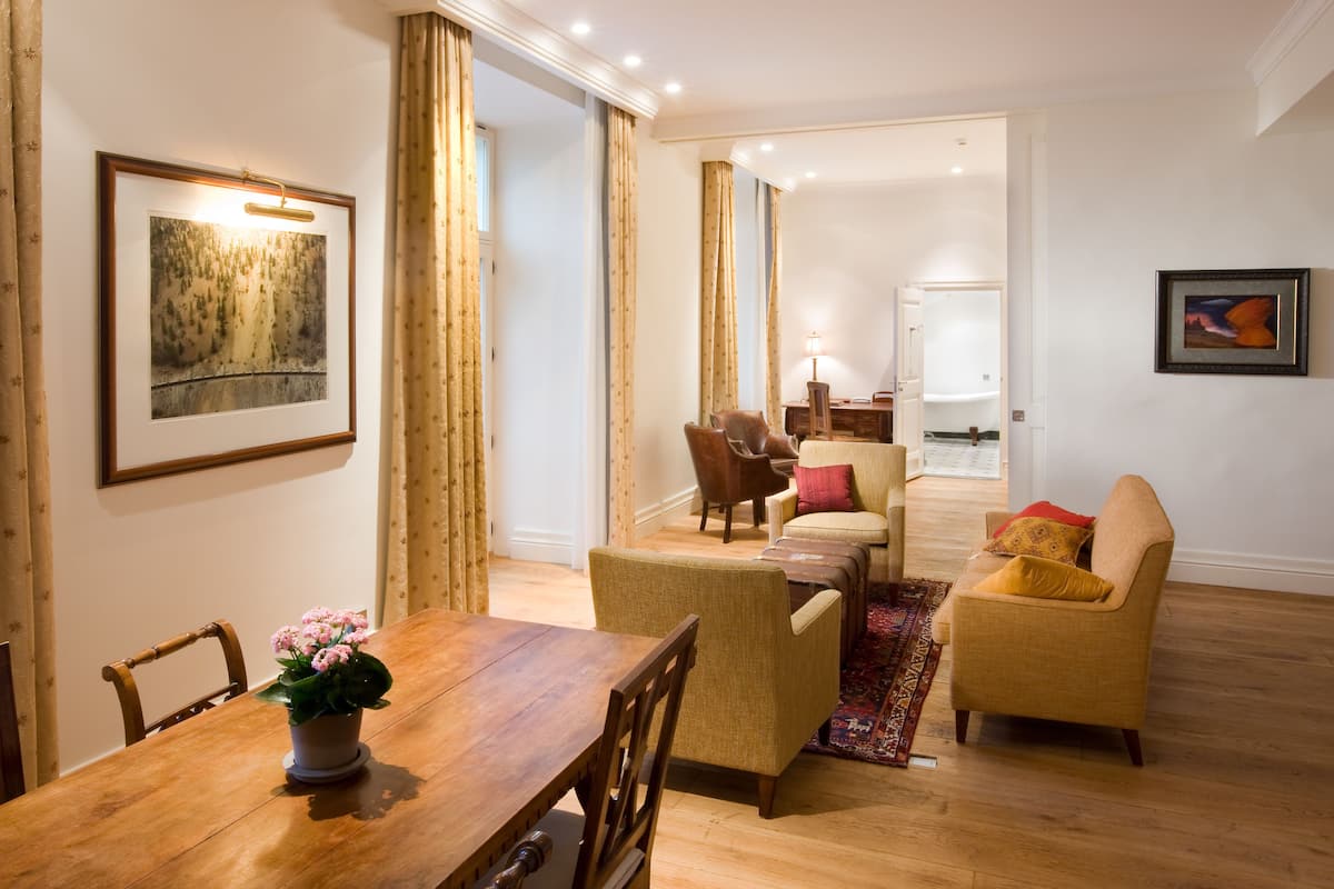 A dining area with a wooden table and chairs, a potted plant, and a view into a living room with a sofa and armchairs.