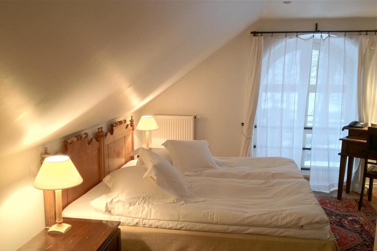 A bedroom with a bed, two bedside lamps, a wooden nightstand, a radiator, a window with sheer curtains, and a desk with a telephone.