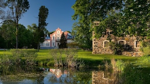 Ein historisches Gebäude mit einer Stein-Fassade und Bogenfenstern, umgeben von üppiger Vegetation und einem Teich.