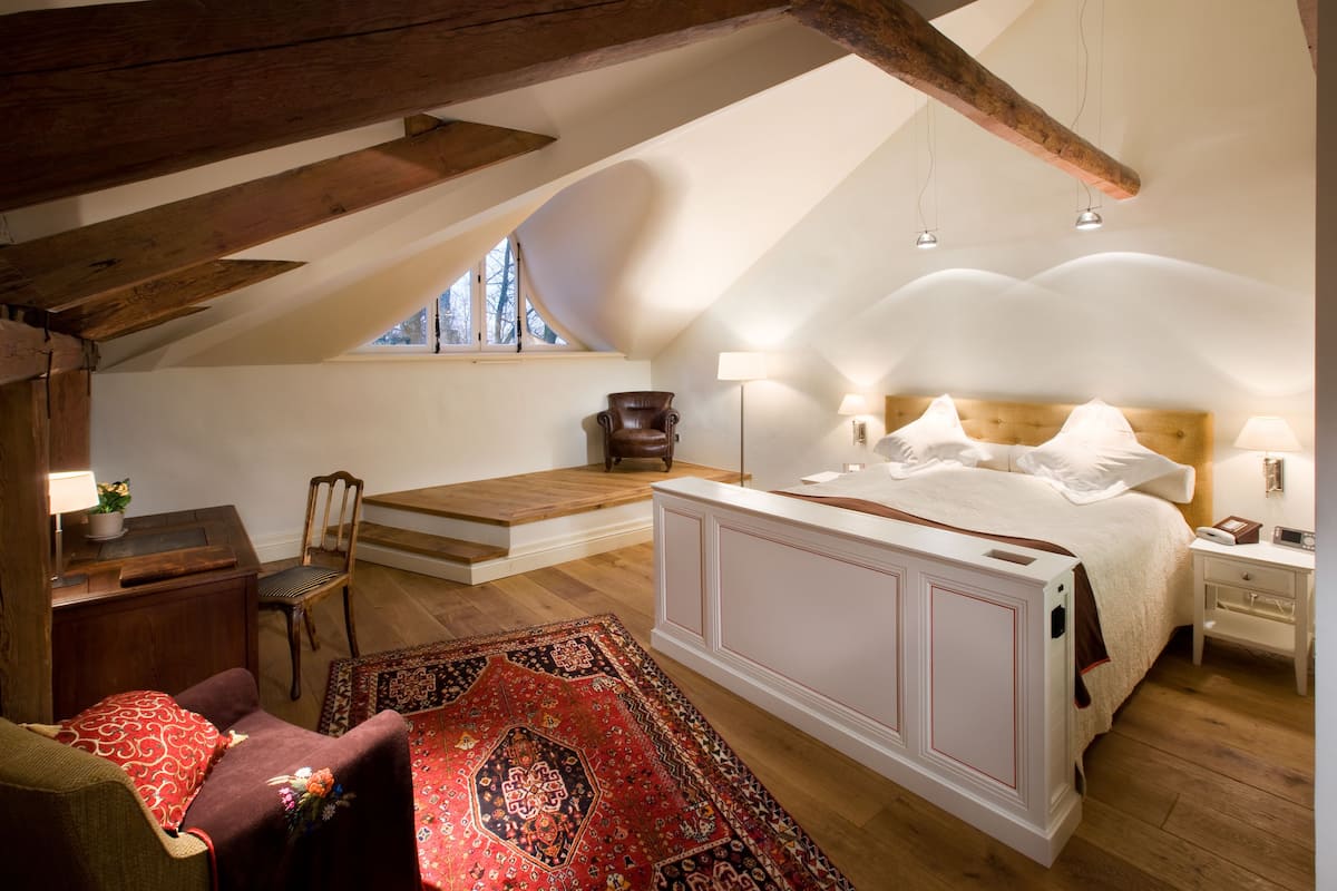 A bedroom with a sloped ceiling, a large bed, a wooden nightstand, a chair, a desk, and a red patterned rug.